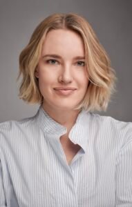 Professional headshot of Cassidy Gard, a woman with short blonde hair wearing a light blue striped shirt. She is facing the camera with a neutral gray background behind her. The lighting is even and studio-style, suitable for author and press use.