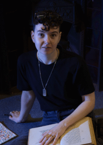 Cassidy Ellis Salter sits on the floor with an open notebook in front of them, looking up at the camera. The room and the light is dark blue.