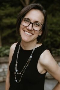 Photo shows a smiling white woman with dark, short hair and glasses. She is pictured from the chest up in a black tank top with a long beaded necklace in front of a short stone wall with green plants above it.