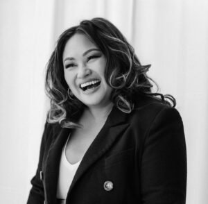 A picture of a Filipino American woman with wavy brown hair and brown eyes. She is shown from neck up, wearing a dark jacket, and is laughing aloud.