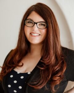Woman with long brown hair wearing cat eye glasses and a navy dress with white polka dots, posed and smiling at the camera.