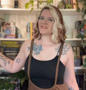 Framed in front of a bookshelf full of plants, books, and nicknacks is a white woman from the waist up. She is wearing clear-framed glasses and has brown hair parted on the side with two blonde strips in the front. She has multiple tattoos on her arms and collarbones/shoulders. She's wearing a black spaghetti strap top with cotton "overall" brown pants.