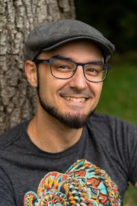 Christopher Roubique, a man with tan skin and a dark chinstrap beard, smiles while seated against a tree. He is wearing a gray newsboy cap and dark blue glasses. We can see only part of his shirt, which is gray with the upper half of a multicolored bear visible.