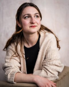 Claire, a white woman with long brown hair, a white cardigan, and a black shirt, stares into the distance over the back of a chair