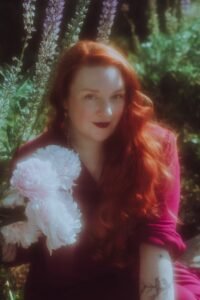 Clare Edge is a white, femme author wearing a dark pink dress. A huge dahlia is next to her face, her hair is very long and red and wavy and she smirks at the camera like she has a secret, which she does.