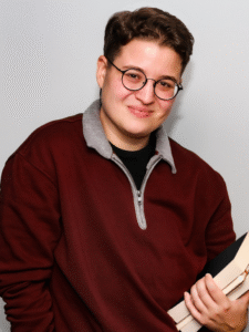 A photo of a person with short, brown hair and glasses, wearing a maroon shirt and holding a stack of books.