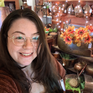 Cynthia Prith, a white woman with wavy auburn hair and round glasses smiling in front of an antique store fireplace covered in copper baking molds and old tea kettles