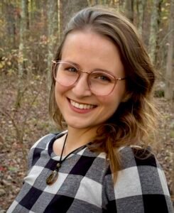 A close up photo of the brown-haired author wearing glasses with pink frames against a fall woodland background.