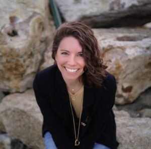 A woman with brown curly wind swept hair smiles at the camera. She is wearing a mustard yellow shirt and black blazer. She is sitting on rocks near the beach.