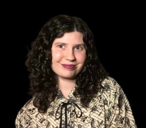 Close up picture of Demri Redmon wearing a tan shirt with a black bow and a black pattern of skulls, ravens, and handwritten text. She’s wearing burgundy lipstick and has short, curly, black hair.