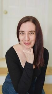 Elayna R. Gallea, a white woman with long brown hair, sits with her head resting on her chin. She is looking at the viewer and has a small smile.