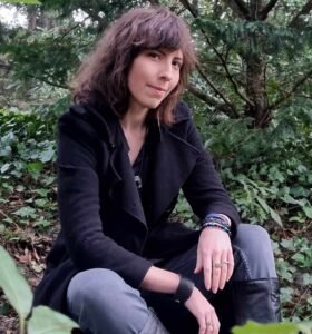 Elian J Morgan, who has dark wavy hair, sits amongst woodland. They are wearing a long black coat, grey jeans, knee-high black boots, and numerous bracelets.