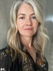 Elizabeth Noel, a white woman with blonde hair in a floral shirt, standing in front of a grey wall.Elizabeth Noel, a white woman with blonde hair in a floral shirt, standing in front of a grey wall.