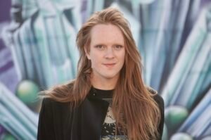 Elle Grenier, a white person with long ginger hair, stands in front of a wall covered with various spray-paint tags. They’re looking at the viewer with a slight smile.
