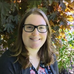 Elysia Rourke, a white woman with brown hair and thick, black glasses, standing outdoors in front of a reddish-brown maple tree. The image is visible from the chest up. She is smiling.