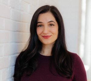 Photo of author Emily Ohanjanians, an Armenian-Canadian woman with long brown hair and brown eyes. She stands against a white brick background, wearing wearing a maroon sweater, and smiling at the camera.