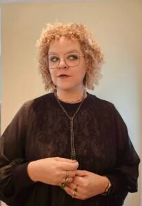 Emma Cole, a biracial woman with short curly hair, stands in front of an off-white wall. She wears a sheer black dress over a dress printed with anatomical hearts and lungs. She is looking up and to the right.