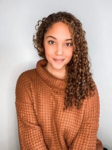 Erynne Rivers, a biracial Black woman with brown curly hair, sits in an orange knit sweater against a white wall backdrop, looking at the camera with a soft smirk.