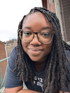 Falencia Jean-Francois, a Black person with shoulder length locs, leans over the railing of their porch, smiling slightly. They are wearing a black t-shirt that says "Tell your dog I say hi" in white letters across the chest.