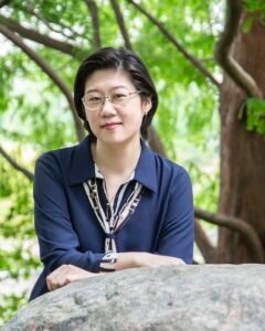 A photo of Florence Chien. She is a mid-30's Chinese woman, with black short hair and a pair of glasses. She is wearing a dark blue shirt and leaning into a boulder set in front of a canopy of trees.