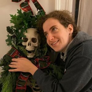 Gillian Daniels, a woman with brown curly hair pulled back messily hugs a plastic skeleton dressed in leaves and a red and green bow. The skeleton is dressed in Christmas decorations. She smiles happily into the distance.