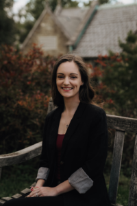 Photo of a white woman with long, brown hair and blue eyes. She is shown smiling in a black blazer and red shirt while sitting on a rustic bench with an autumn garden and cottage behind her.