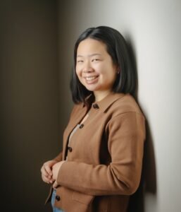 Gracie Marsden, an Asian woman with shoulder length hair, leans against a wall, smiling mischievously at the camera.