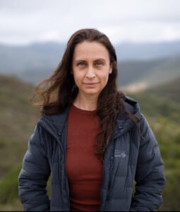 A white woman with long dark hair stands in the mountains looking straigh at the camera. She wears a rust-colored shirt and black puffy jacket. She has a slight smile.