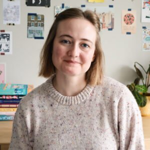Hannah Brohm, a white woman with a blonde bob, sits in front of a wall with postcards and book stacks. She is looking at the camera with a slight smile.