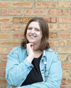 Harper Kinsley, a young, white woman with medium-length, brown hair smiles while leaning against a on orange and brown brick wall. She wears a denim jacket over a black top.