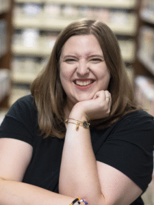 A woman with shoulder length, dark blonde hair smiles at a camera, resting her chin in one hand. She is sitting between two book shelves and wearing gold bracelets.