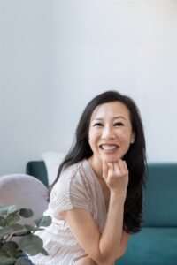 Heather Eng, the author of Double Happiness, sits with her chin resting on her hand. She is an Asian American woman in her early 40s with long, slightly wavy black hair. She is looking directly into the camera and smiling widely.