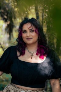A woman with brown and pink hair wearing a black top. Background: green foliage.