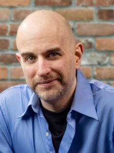 Photo of J.A. Morgenstein, a bald white man with a short beard, posing in front of a brick wall.