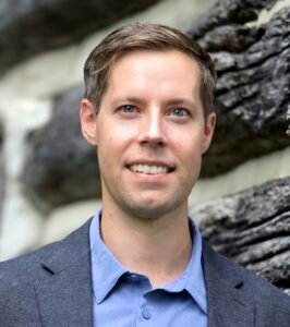 J.J. Kochmanski, a white man with short sandy hair and blue eyes, standing outside a rustic farmhouse. Dressed in a blue shirt and heather navy blazer, he is looking at the viewer and smiling.