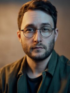 J.J. Unthank, a white man with stubble and short brown hair, wearing round glasses and a green jacket.