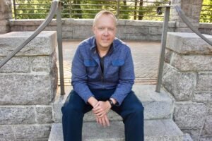 A picture of a white male in his mid-forties with blonde short hair and blue eyes. He is shown seated on a gray cement stair and is wearing a blue zip-up sweater, black t-shirt, and blue slacks.