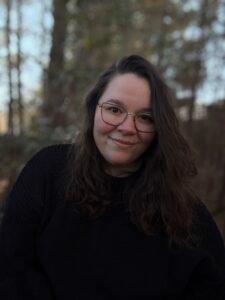Image of Jenee, a white woman with brown hair, bust up in a black sweater with trees behind her.