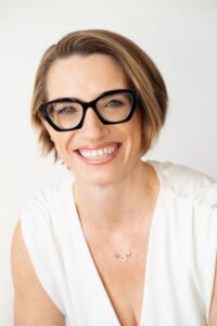 A white woman in a white sleevless shirt, short brown hair, big glasses. She is smiling.