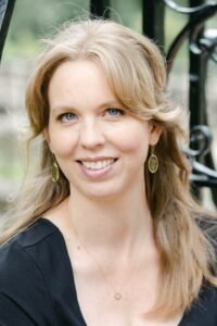 Jennifer Mandula, a white woman with long blond hair and bangs, is smiling happily at the camera. She is wearing a black dress, and there is some leafy greenery in the background.