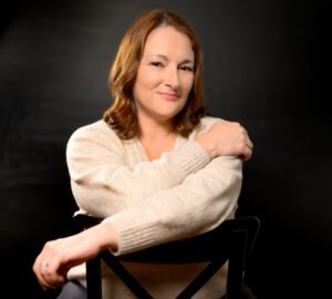 A Caucasian woman sitting backwards on a black chair with her arms crossed. She has reddish brown hair and brown eyes and is smiling at the camera. She is wearing a beige/cream colored sweater.