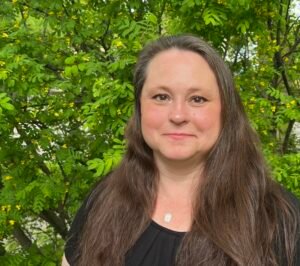 Photo shows a white woman with long, dark hair and brown eyes. She looks directly into the camera with a slight smile on her face.