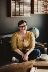 Photo of Jessi Cole Jackson, a white woman sitting cross-legged on a couch, smiling.