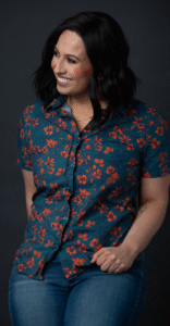 Photo of Jesse Aragon, wearing blue jeans and a dark turquoise shirt with orange flowers. She is looking off to the side and smiling.
