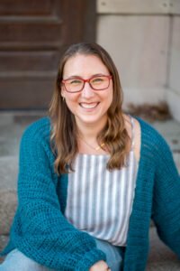 Jessica M. Felleman, a white woman with medium length light brown hair and blue eyes, wearing red rectangle framed glasses, and sitting outside on stone steps leading to a brown door. She is wearing a vertically striped grey and white shirt and a sea-blue crocheted cardigan and blue jeans. She is looking at the viewer and smiling.