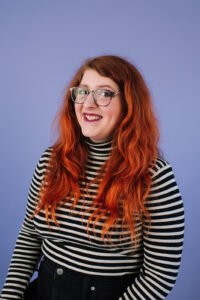 Jessica Fowler is smiling with red hair in a black and white striped shirt with a purple background.