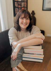 Jess Mey, a white woman with shoulder-length dark brown hair sits on a bench, smiling, with her arms over a pile of books on a table, spines turned away.
