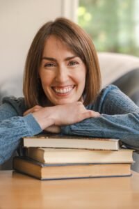 Jillian Laine, a white woman with medium length light brown hair sits with a stack of books. She is smiling.