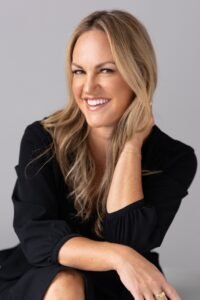 Joan F. Smith, a white woman with long brownish blonde hair, sitting with her arms folded. She's wearing a black top and has her arm tucked behind her head. She's smiling.