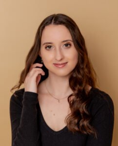 Julia smiles softly at the camera with her hand by her face. Her brown hair curls slightly and she wears a black long-sleeved shirt with a small moon necklace.